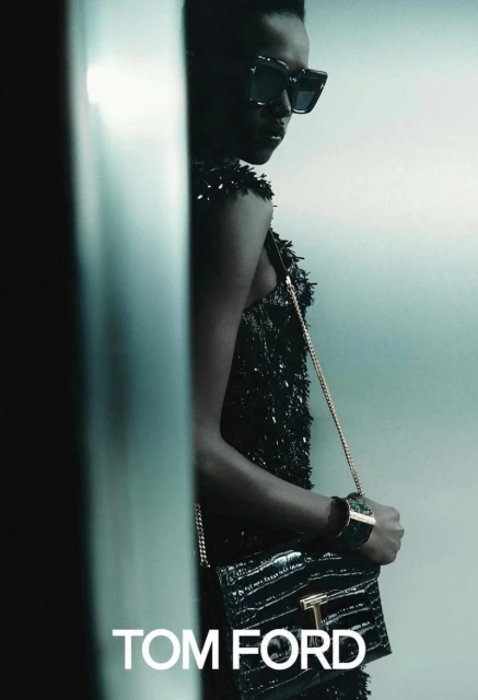 Fashion shot of an African woman wearing a black dress, handbag, and sunglasses, posing indoors with a metallic-looking background and Tom Ford text.