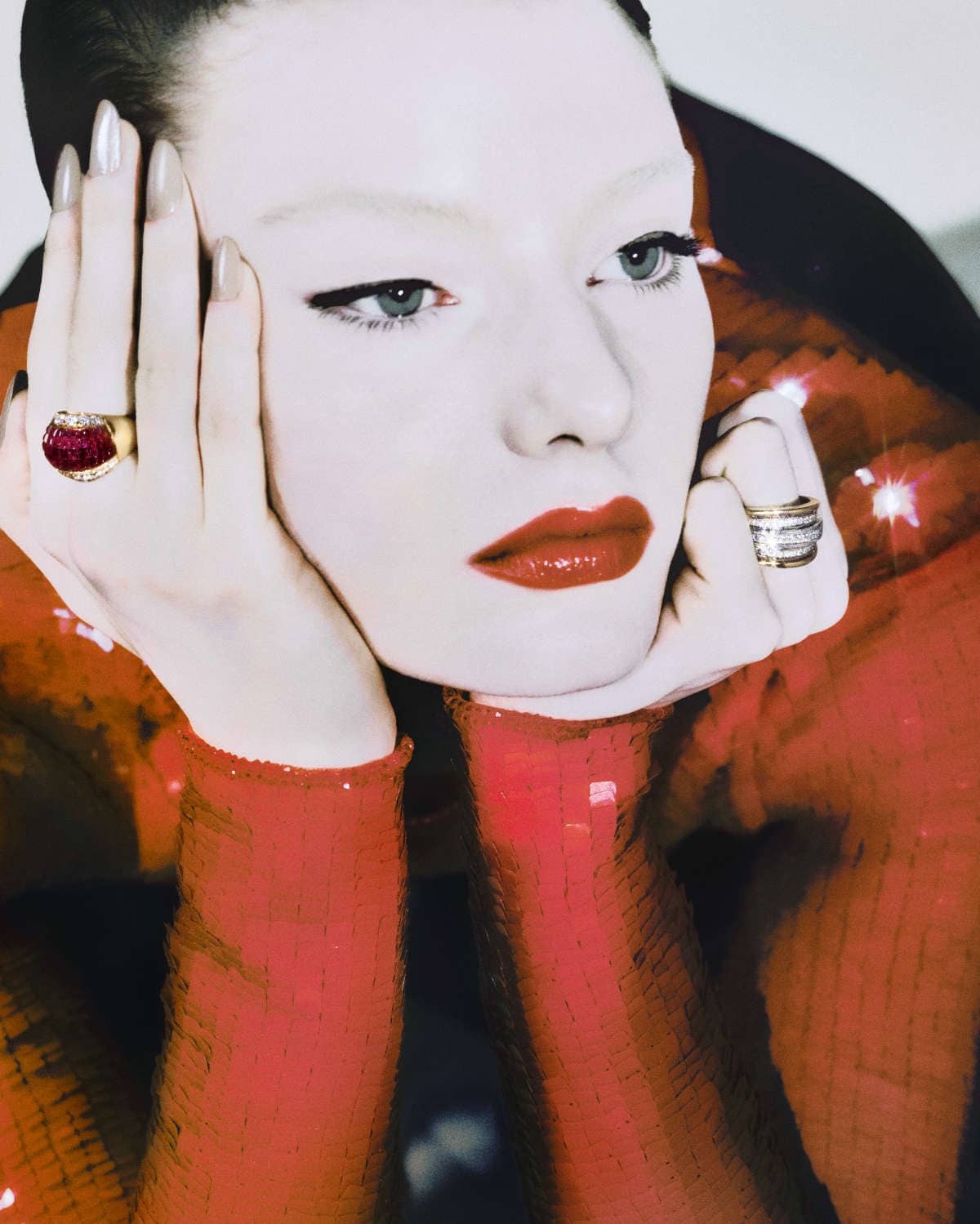 Close-up of a Caucasian woman with pale skin and red lipstick, wearing statement rings, set against a blurred red sequin backdrop.