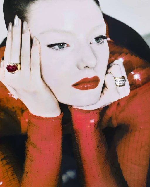 Close-up of a Caucasian woman with pale skin and red lipstick, wearing statement rings, set against a blurred red sequin backdrop.