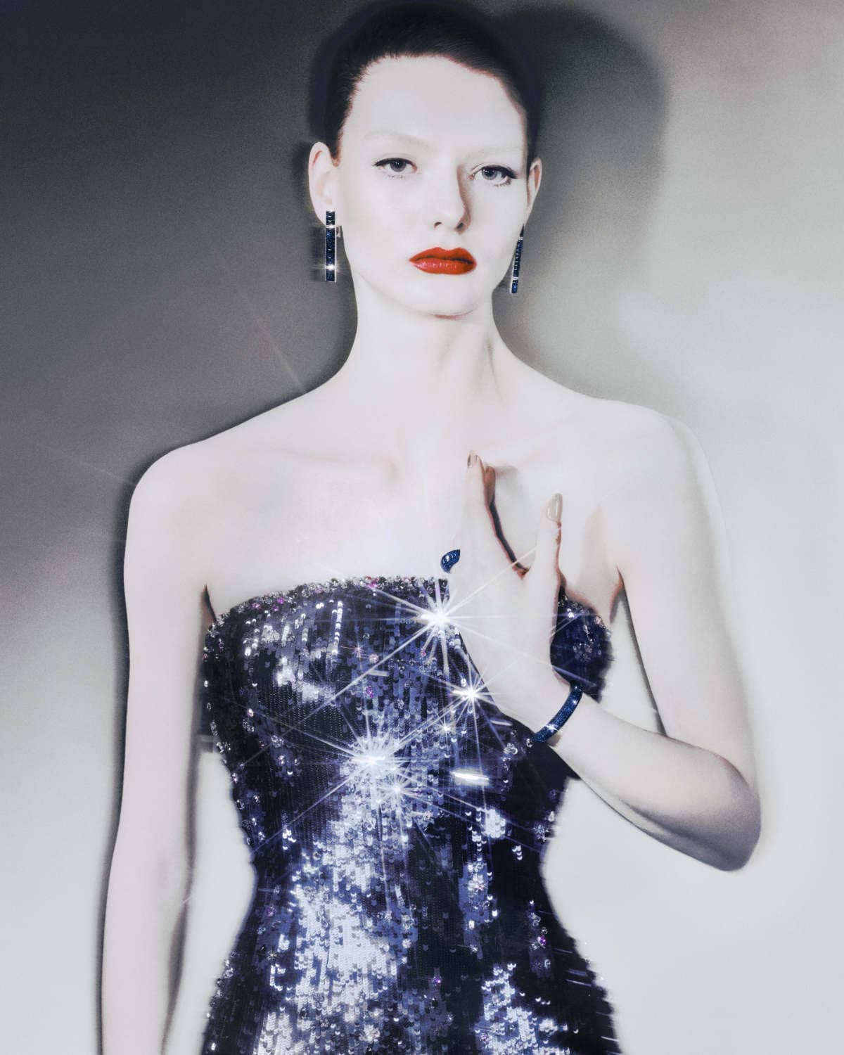 Glamorous woman in a strapless sequin dress, wearing jewelry, posing against a muted gray backdrop. Fashion, beauty, and studio portrait.