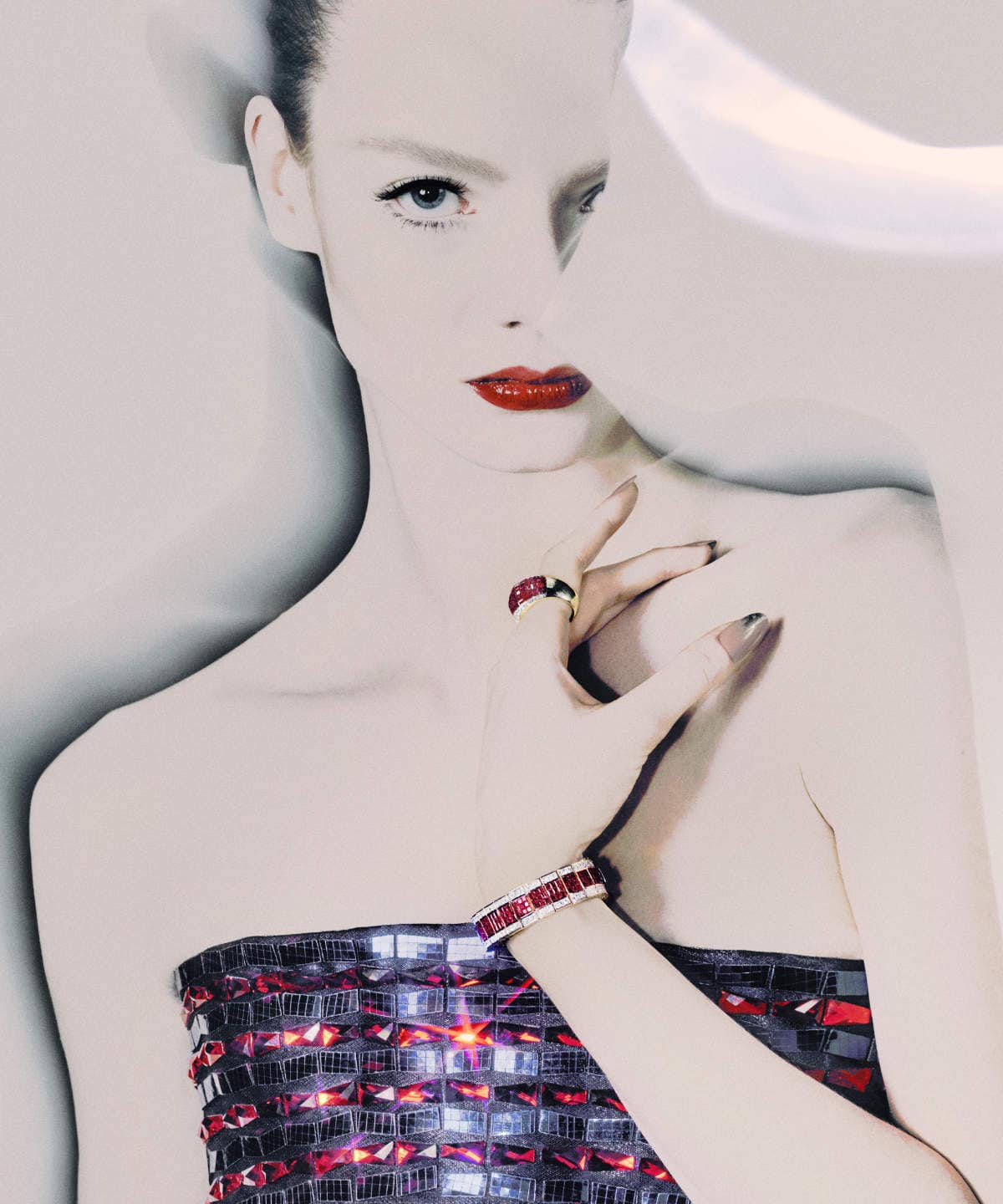Close-up of a fair-skinned woman in a strapless jeweled top with red lips, wearing a matching bracelet and ring against a light background.