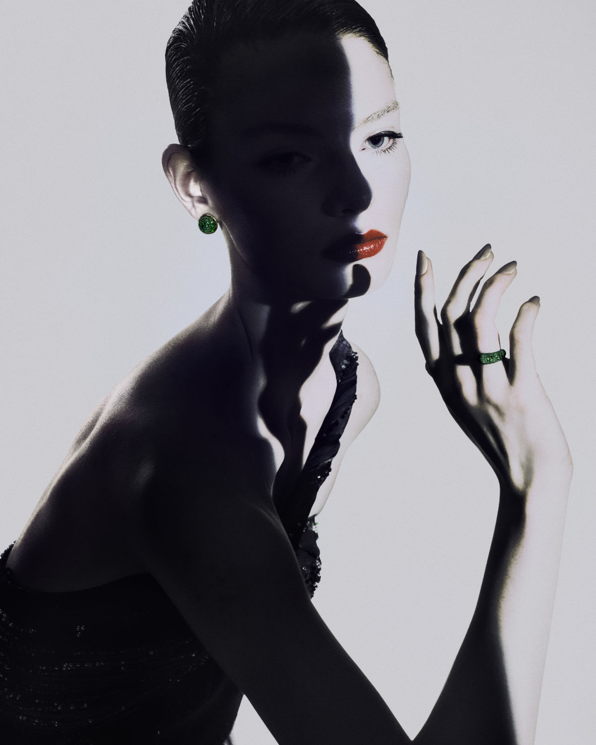 Close-up portrait of a woman wearing dark clothing, green earrings, and a ring, with half of her face shadowed against a white background.