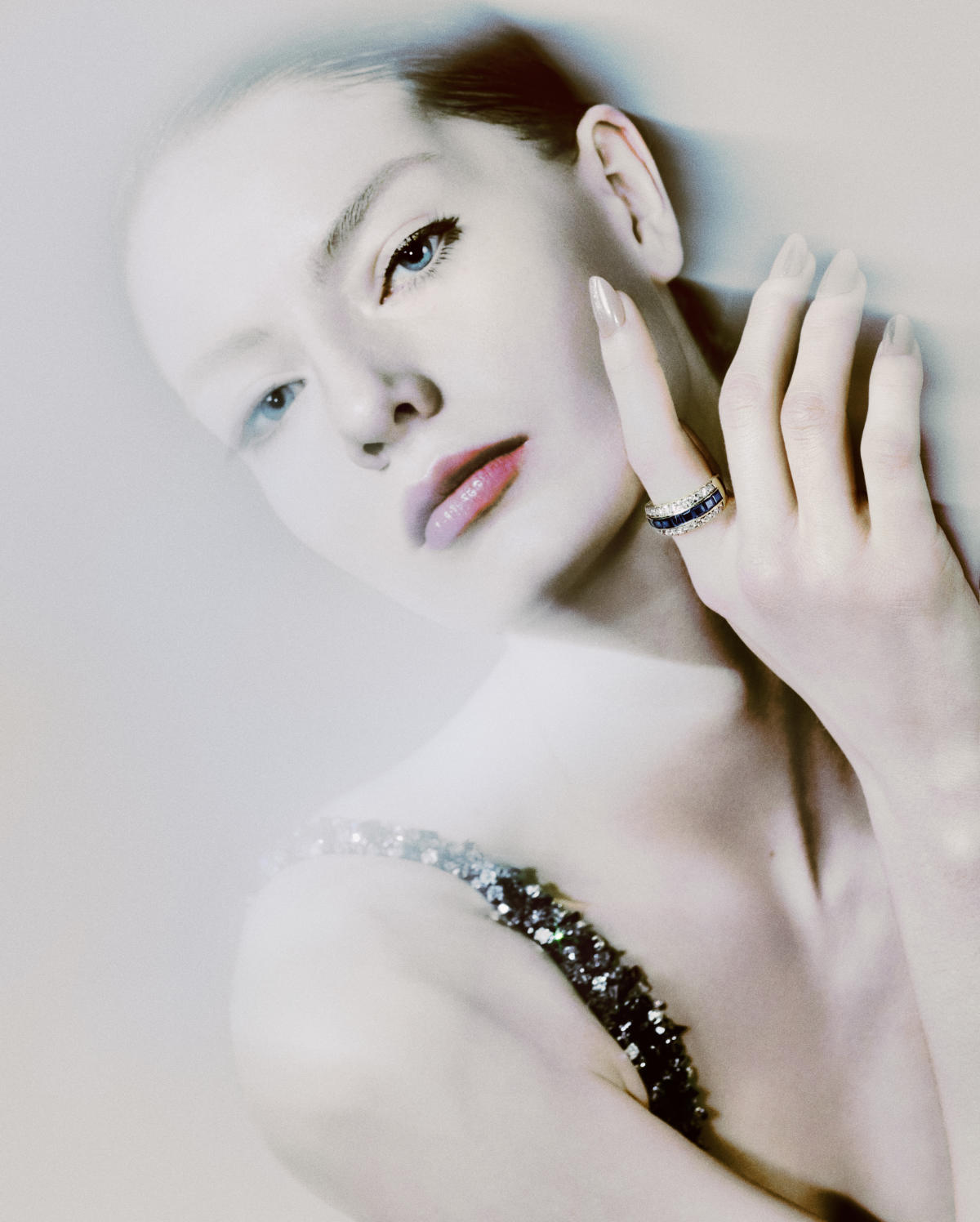 Close-up of a woman with fair skin and blue eyes, wearing a ring and a glittery top against a bright background.