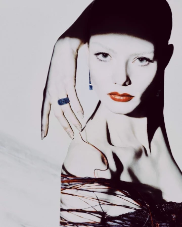 Portrait of a woman with hand on her head wearing blue ring and blue earring with red lips against a textured white background.