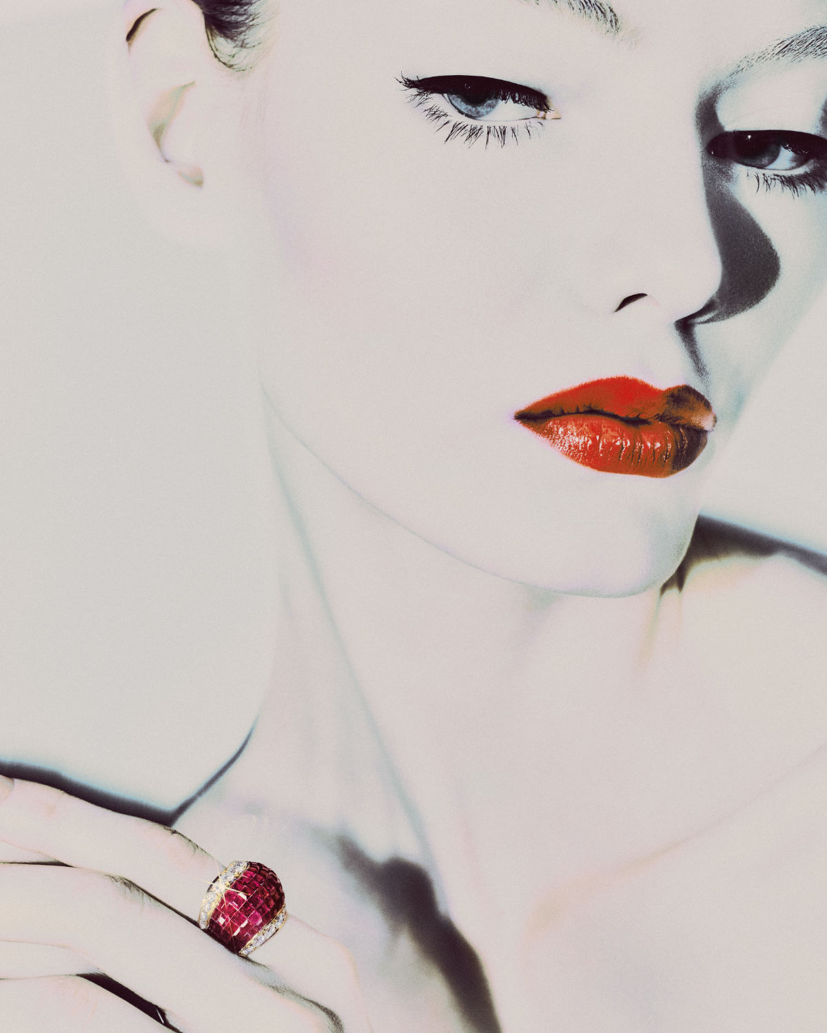 Close-up portrait of a fair-skinned adult female model with vivid red lipstick and a large ruby and diamond ring in a studio setting.
