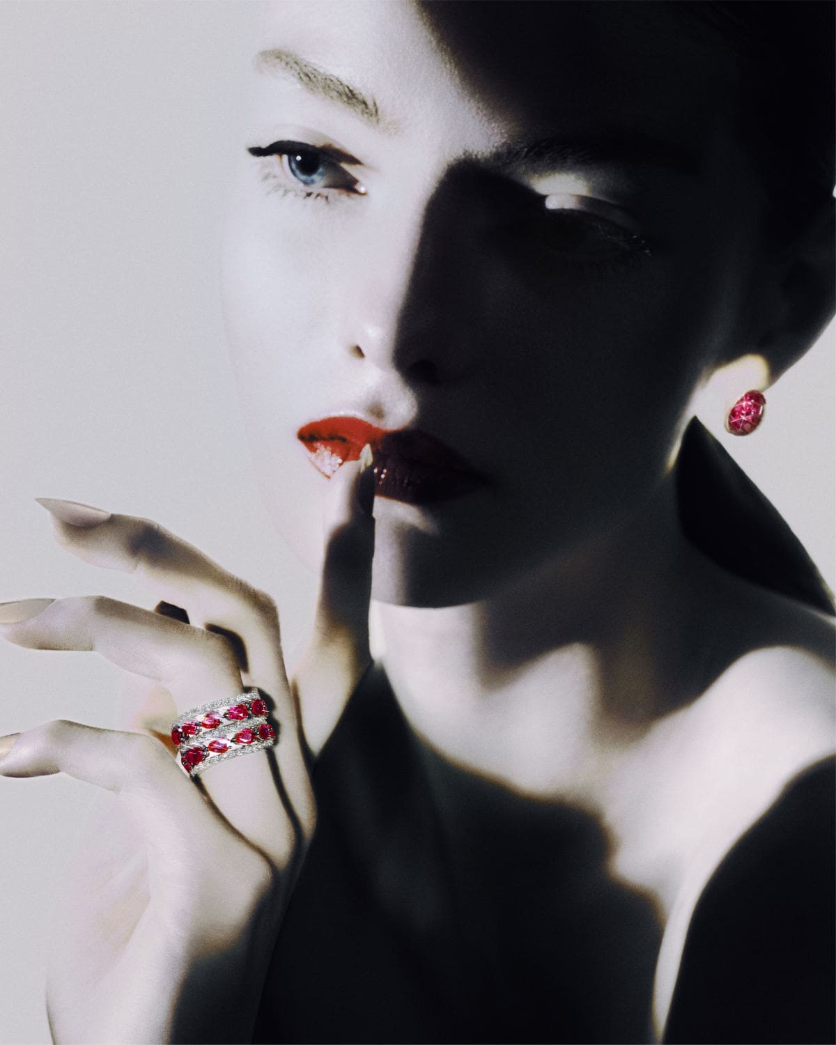 A young adult woman with red lips and blue eyes poses with jewelry, wearing earrings and a ring against a light background in studio light.