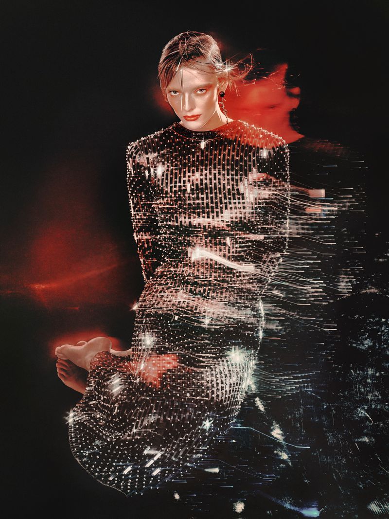 An adult female fashion model poses indoors wearing a glittering black dress, against a dark background with blurred red light effect.