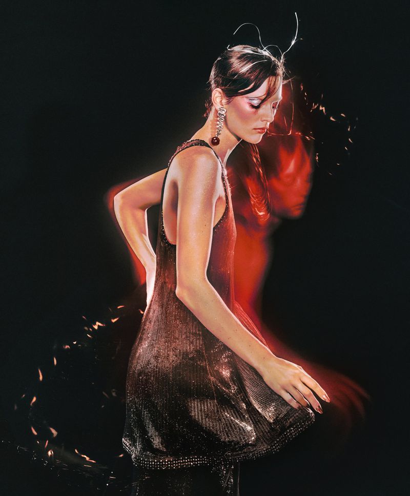 Elegant adult female in a shimmering short dress with red earrings, hands on her hips, artistic lighting, dark background.