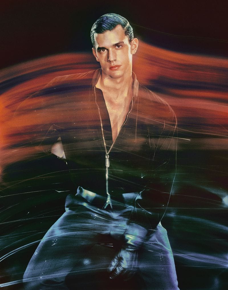 Portrait of an adult man in a black zip-up shirt and gloves, kneeling against a dark background with streaks of color and light.