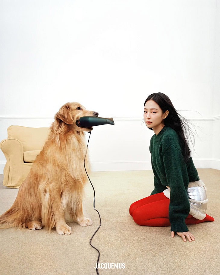 Woman kneeling next to a Golden Retriever with a hair dryer in its mouth, indoors, wearing a green sweater, red tights, and silver shorts.