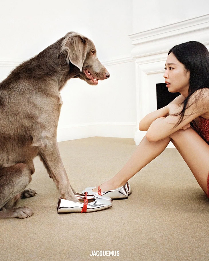 A woman in a sparkling dress sits near a Weimaraner dog with its paw on a pair of silver shoes, in a minimalist setting with the brand logo.