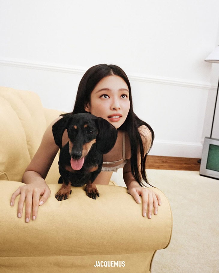 A young woman sits on a yellow couch next to a Dachshund dog; low-angle, eye-level shot with a white wall and minimal decor in background.