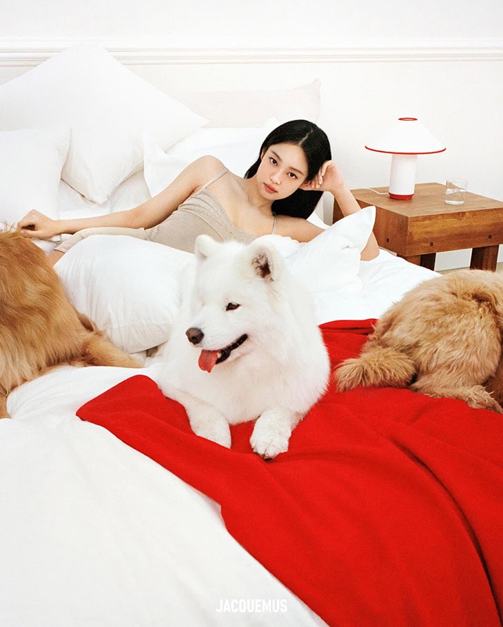 A woman in a beige dress poses on a bed with three dogs, pillows, and a red blanket, next to a wooden table with a lamp.
