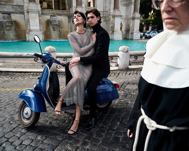 Fashion shot of a couple posing on a moped in front of a fountain, with a nun walking past them. Editorial fashion, outdoor lifestyle.