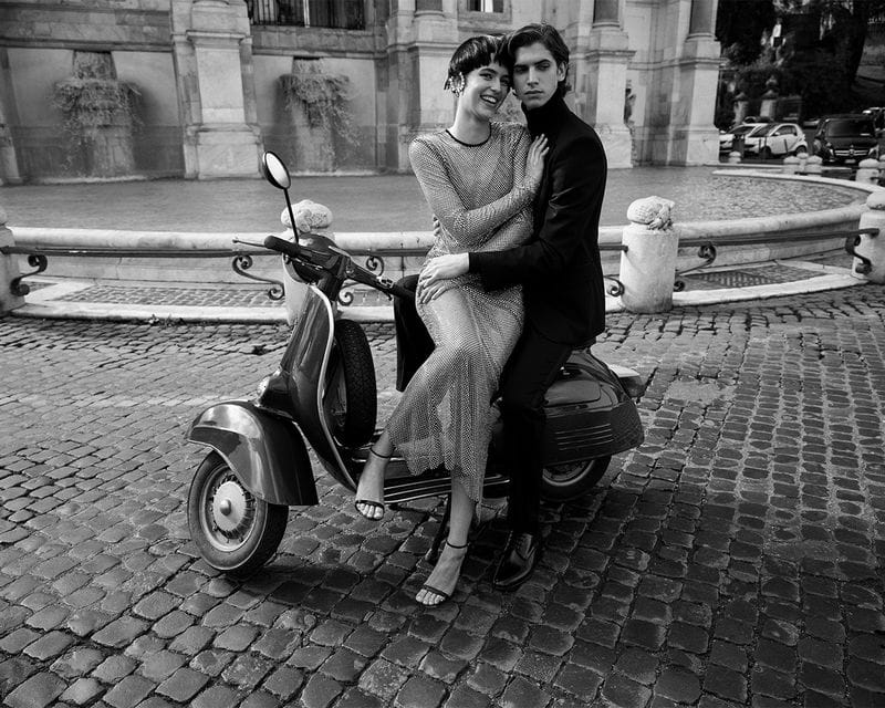 Monochrome fashion shot of a couple sitting on a vintage scooter in Rome, Italy, with a classic fountain in the background on cobblestones.