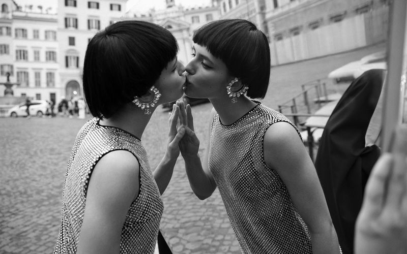 Two women in sparkling dresses appear as mirror images in a grayscale outdoor shot in a European city, showcasing fashion and style.