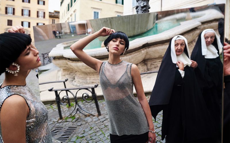 Fashion shot with a young woman in a silver top holding a photo of two nuns, set against a backdrop of a fountain in Italy.