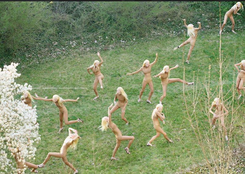 Aerial view of group of fair-skinned female adult dancers in tan bodysuits and blonde hair on a grassy lawn performing choreography.