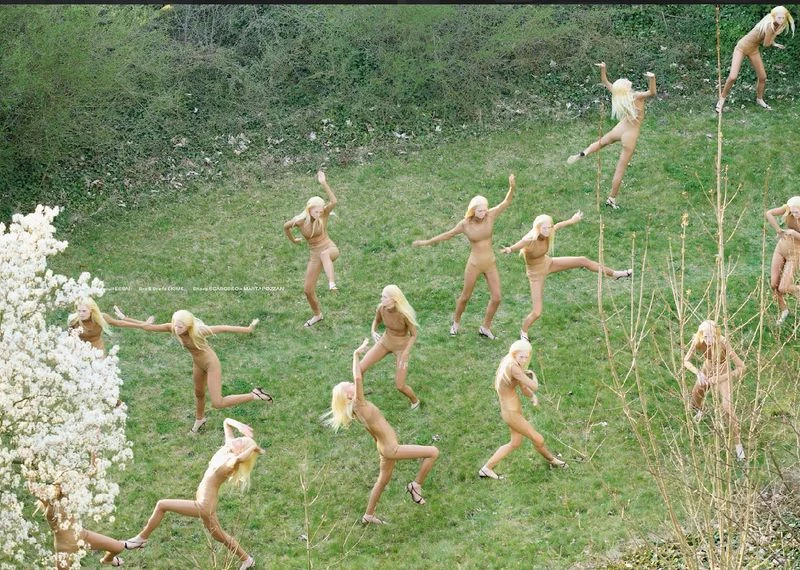 Aerial view of group of fair-skinned female adult dancers in tan bodysuits and blonde hair on a grassy lawn performing choreography.