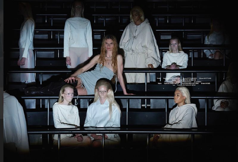 Fashion show presentation featuring a woman in a dress posing with seated women wearing matching white outfits in a tiered seating environment.