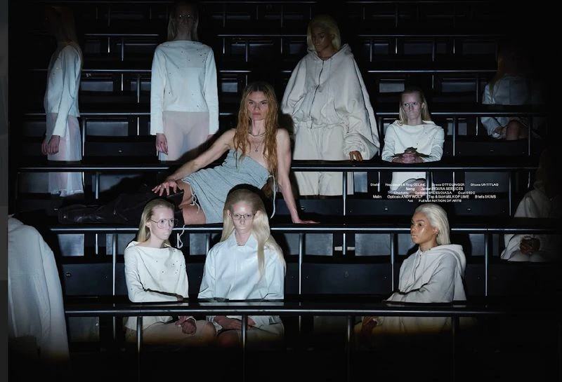 Fashion show presentation featuring a woman in a dress posing with seated women wearing matching white outfits in a tiered seating environment.