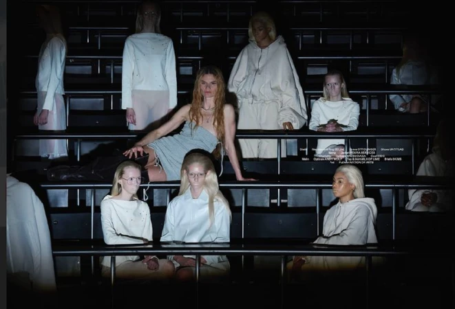 Fashion show presentation featuring a woman in a dress posing with seated women wearing matching white outfits in a tiered seating environment.