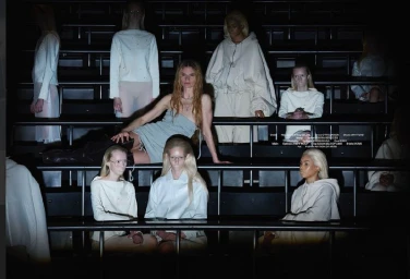 Group of models in white and grey apparel seated on dark tiered bleacher seating in a dim interior environment.