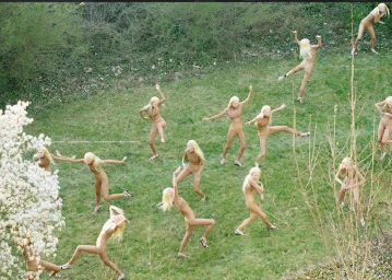 Multiple identical blonde women in nude bodysuits dancing across a green grassy field next to flowering trees.