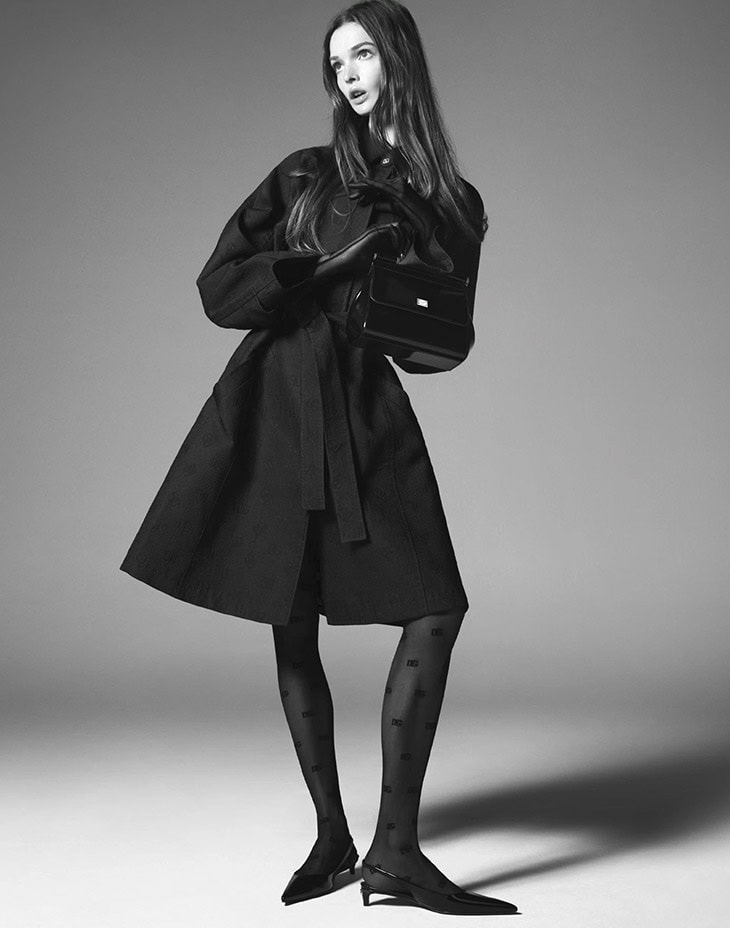 Elegant woman in black coat, patterned tights, and kitten heels holds a structured black handbag against a neutral grey backdrop.