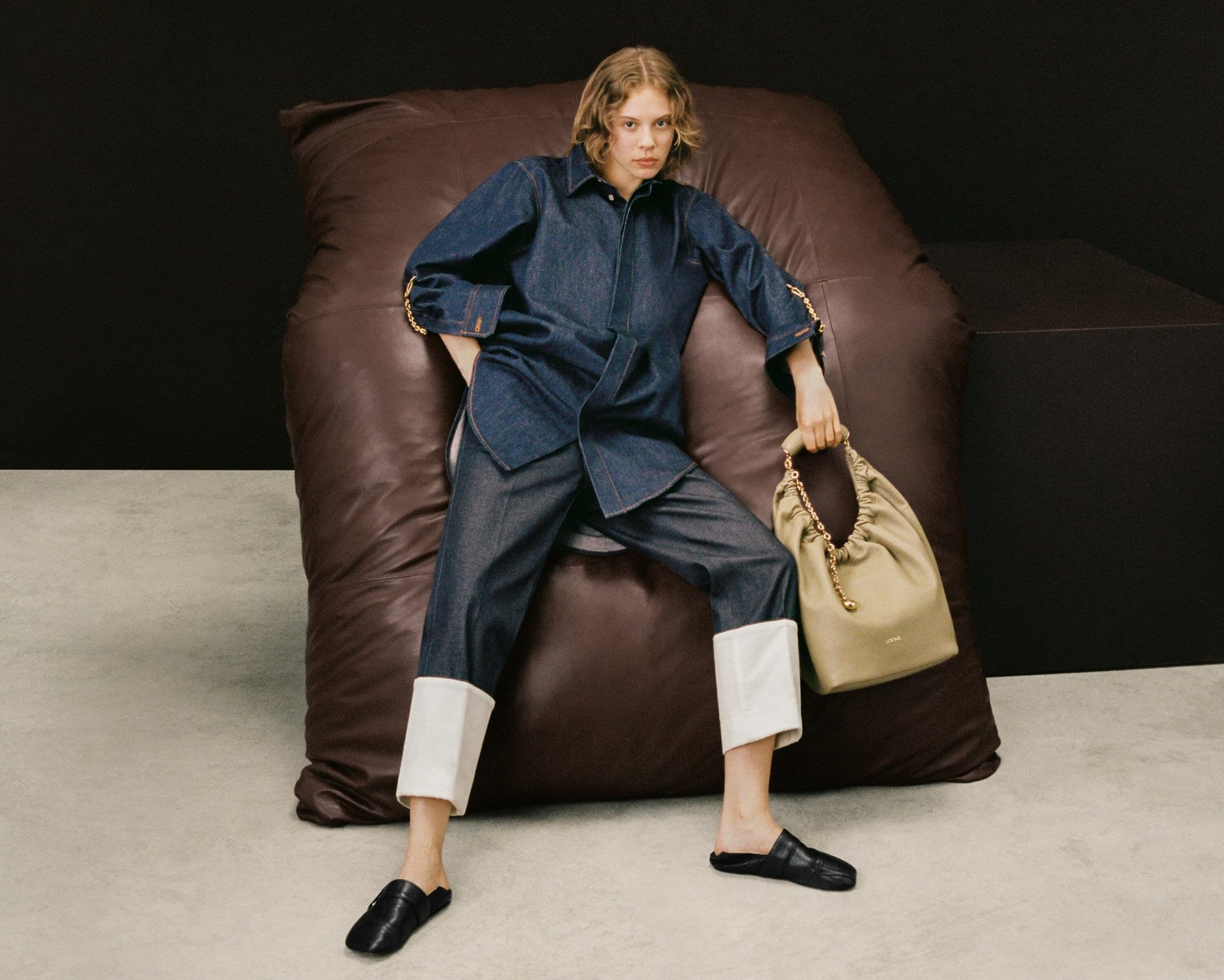 A fashion shot of a young woman in a denim outfit holding a tan purse, seated on a brown leather cushion against a dark backdrop.