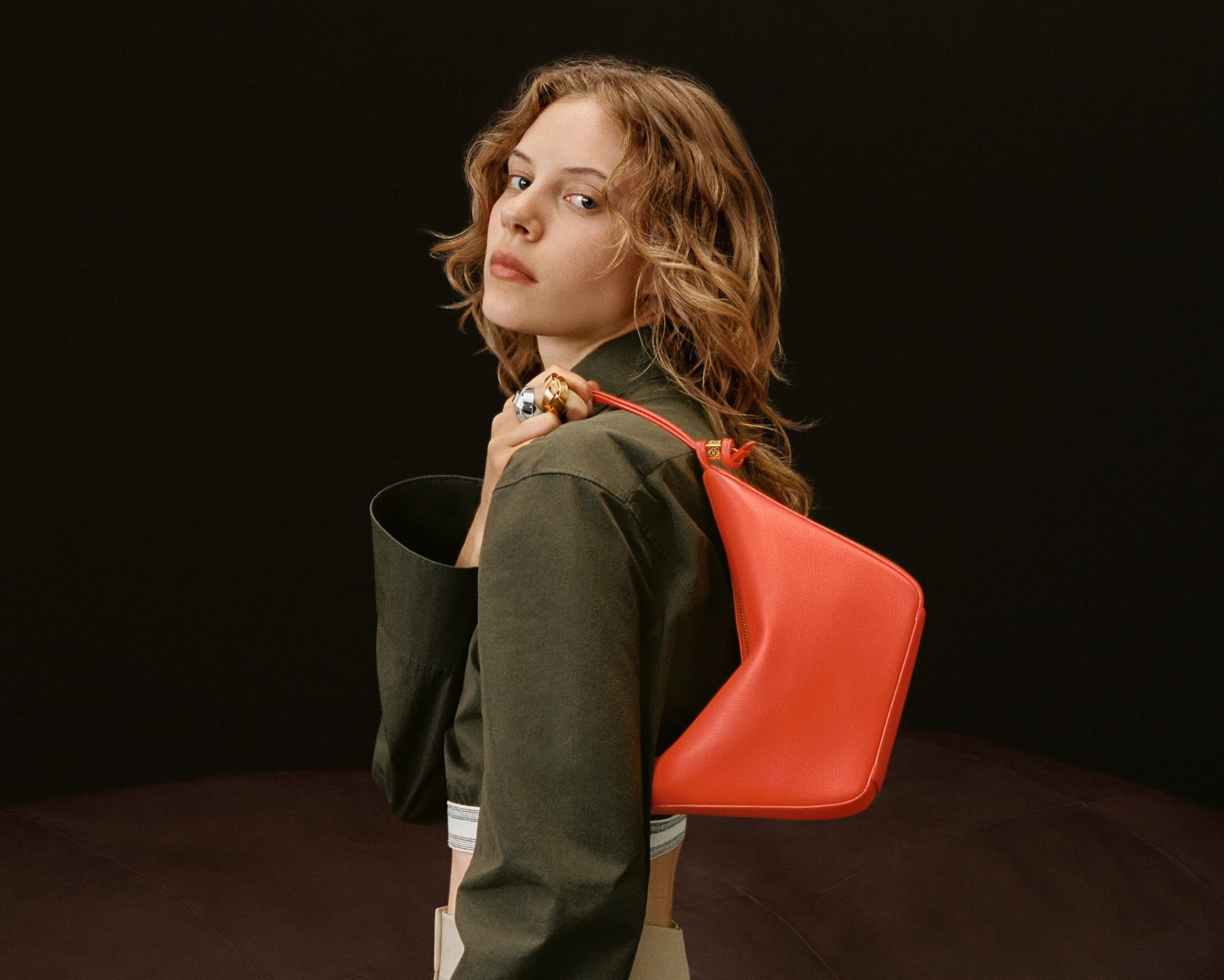 A fair-skinned woman with wavy blonde hair poses in a studio. She wears an olive jacket and carries a coral-colored triangular bag on her shoulder.