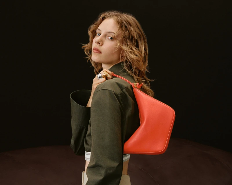 A fair-skinned woman with wavy blonde hair poses in a studio. She wears an olive jacket and carries a coral-colored triangular bag on her shoulder.