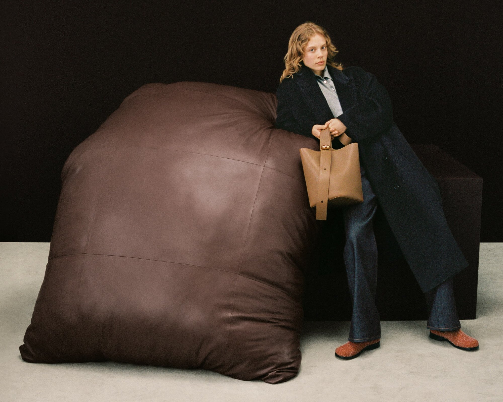 Adult female model in a studio leans on a large brown leather beanbag chair, holding a tan leather bucket bag, wearing a dark coat.