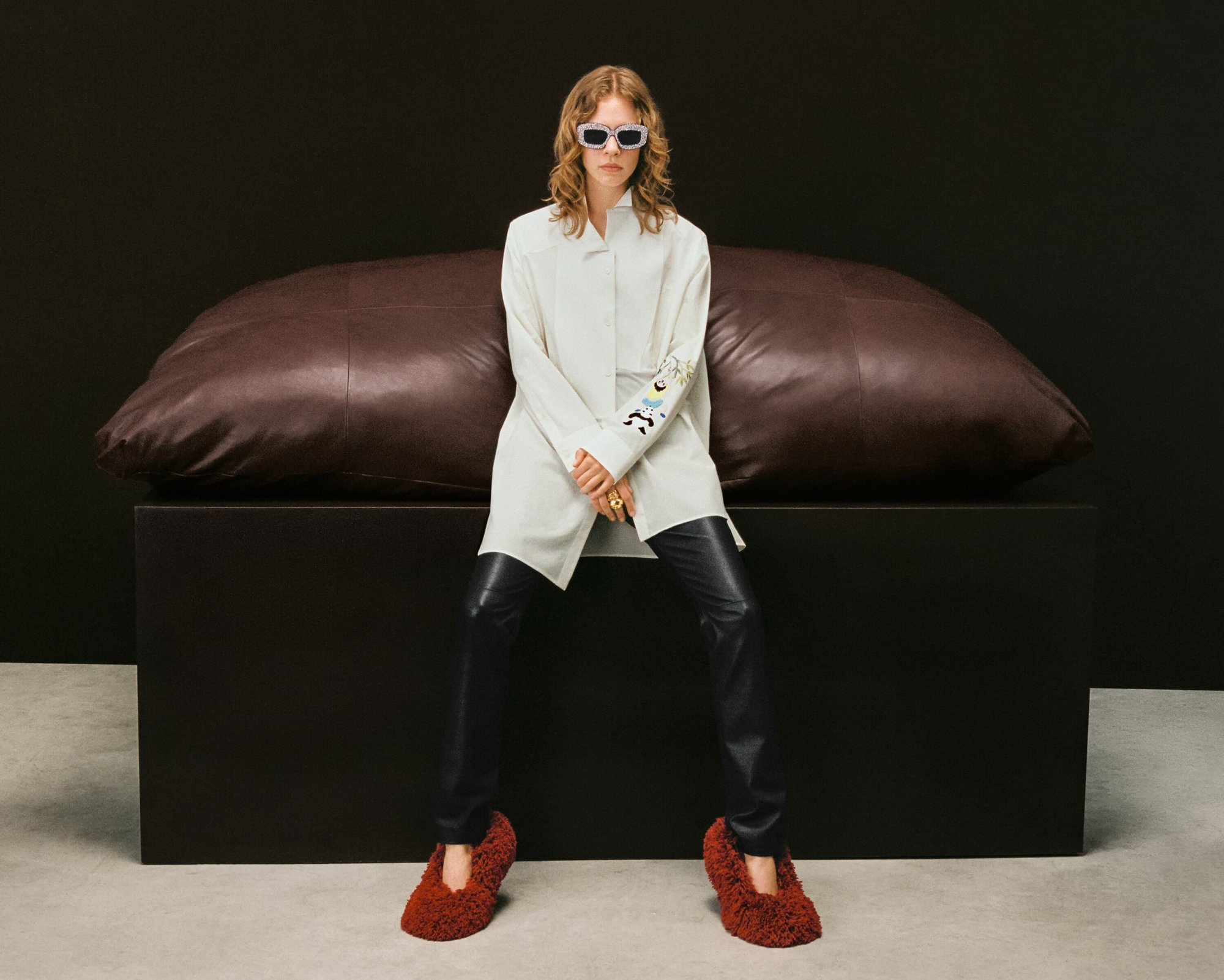 Fashion model sits on a large pillow in a studio wearing a light-colored coat, black pants, sunglasses, and fuzzy slippers.