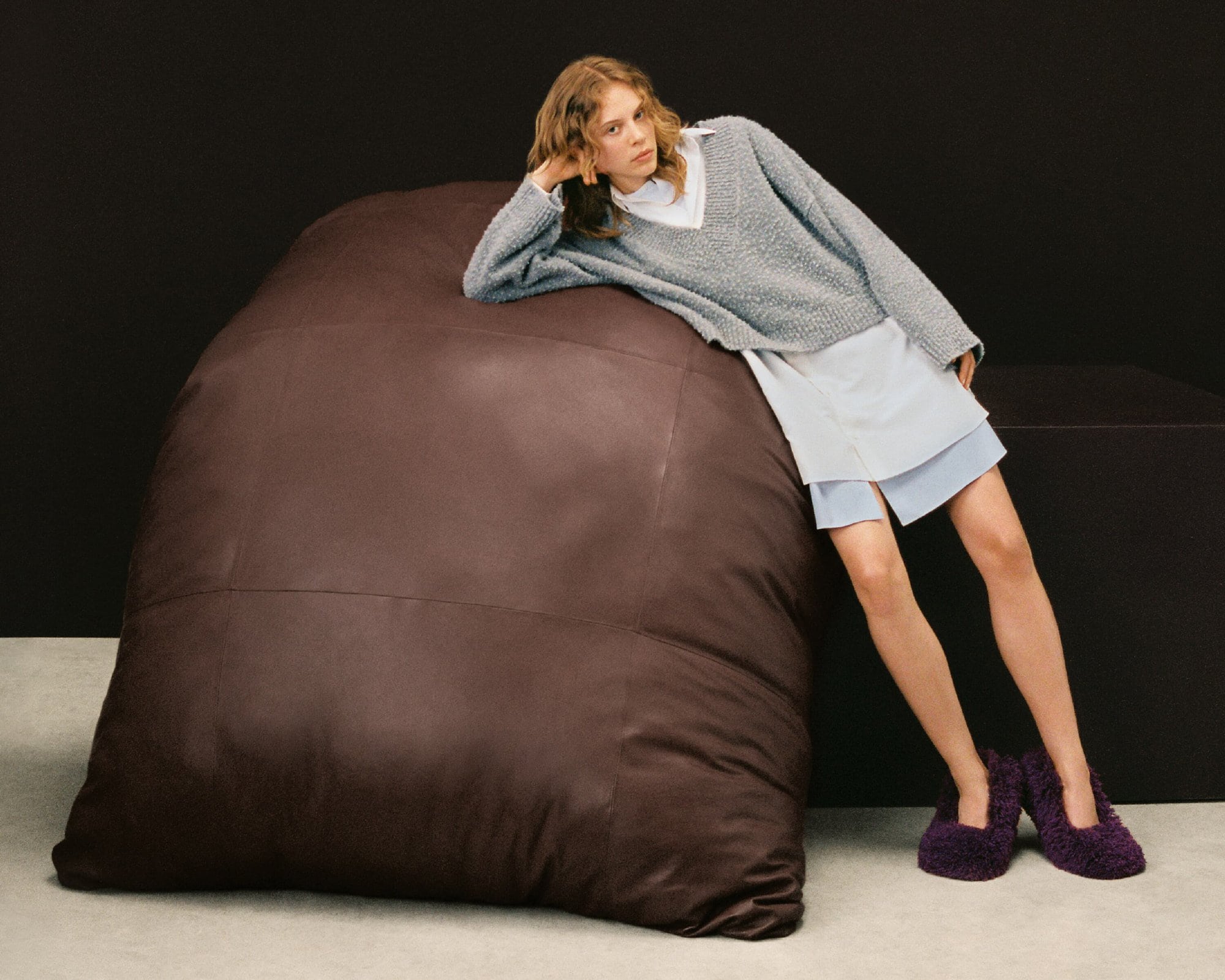 Woman in oversized knitwear and slippers rests on a large leather beanbag in a minimalist setting against a dark backdrop.