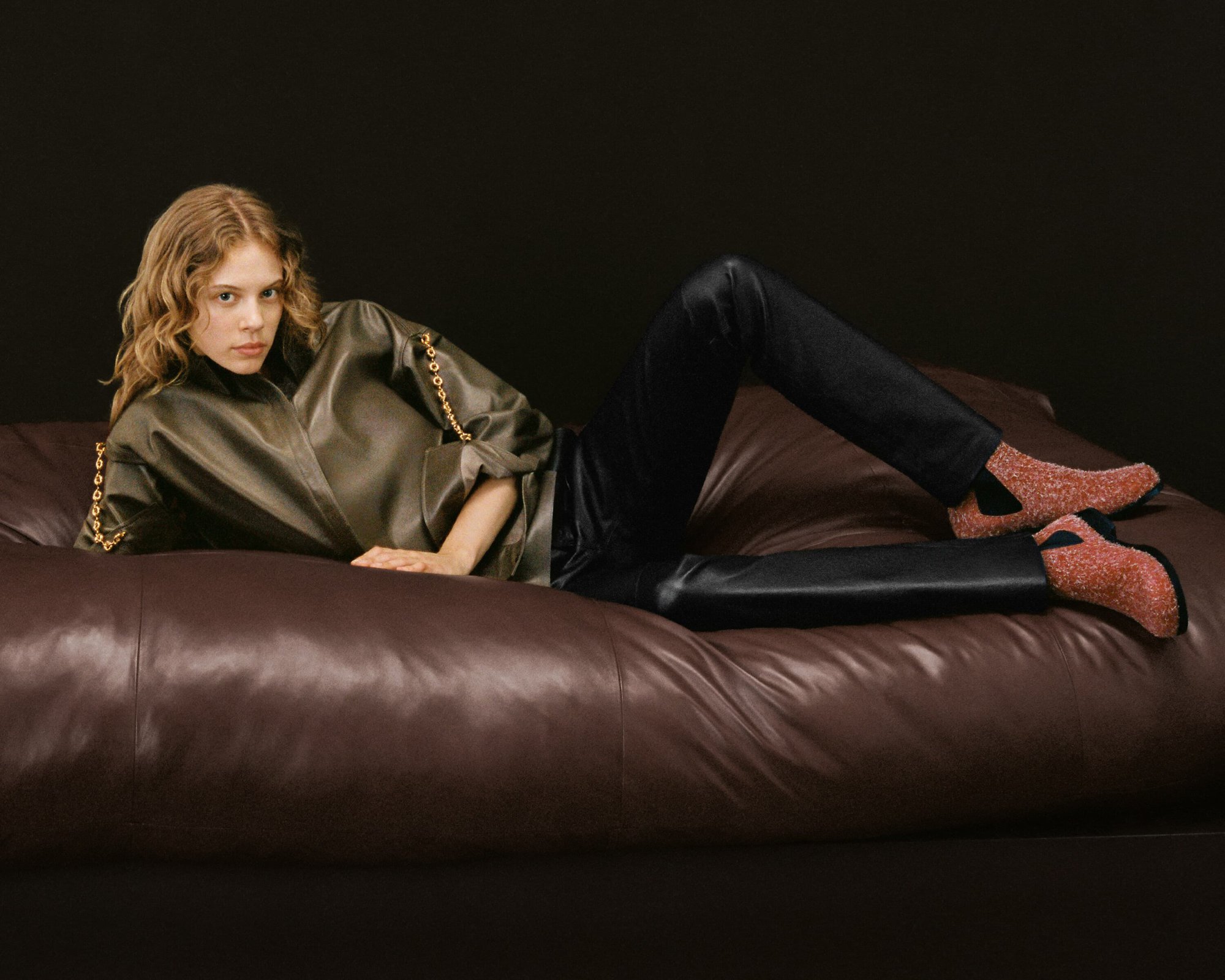 A fair-skinned woman with wavy blonde hair is reclining on a brown leather couch wearing a green jacket, black pants, and pink shoes.