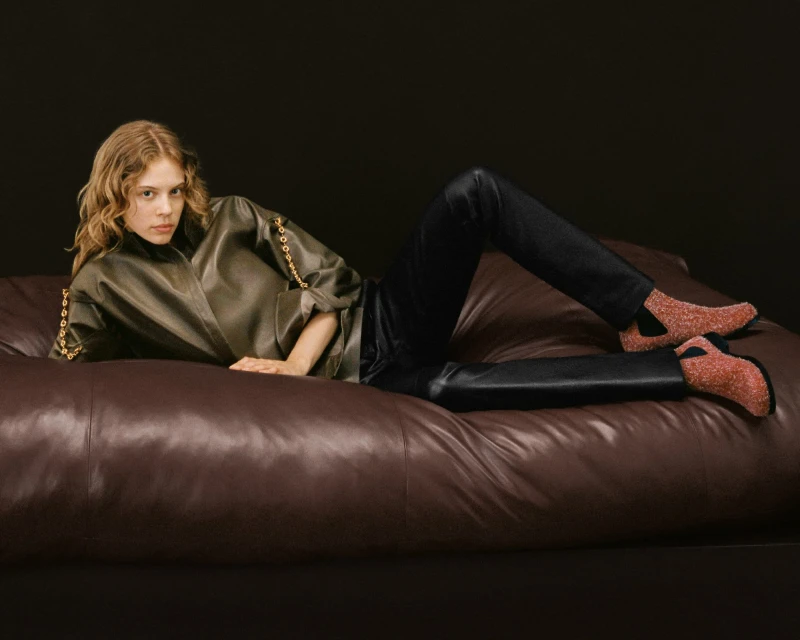 A fair-skinned woman with wavy blonde hair is reclining on a brown leather couch wearing a green jacket, black pants, and pink shoes.