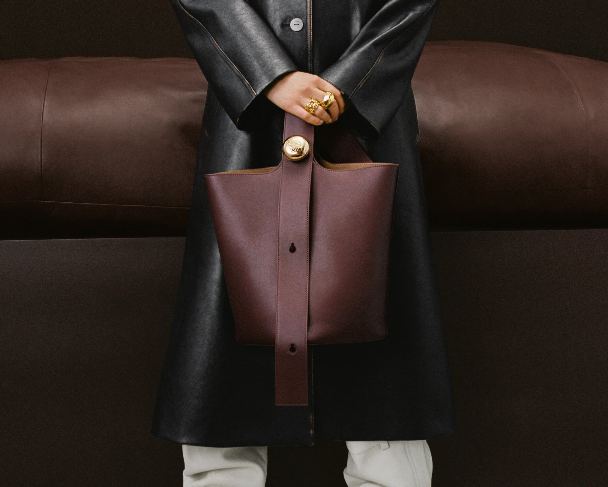 Close-up of a person holding a structured dark burgundy bucket bag in front of a black leather coat against a dark brown background.