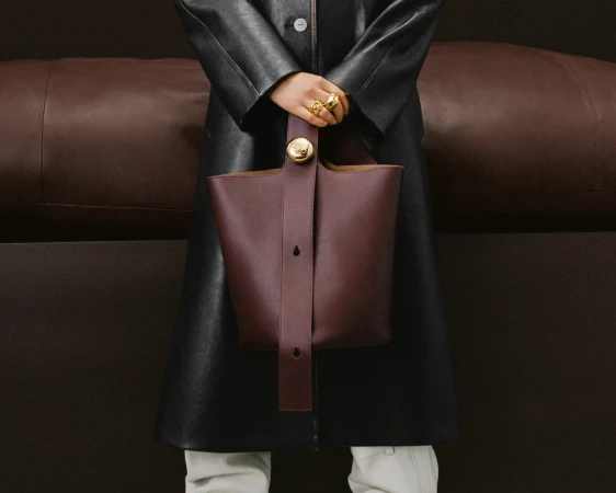 Close-up of a person holding a structured dark burgundy bucket bag in front of a black leather coat against a dark brown background.