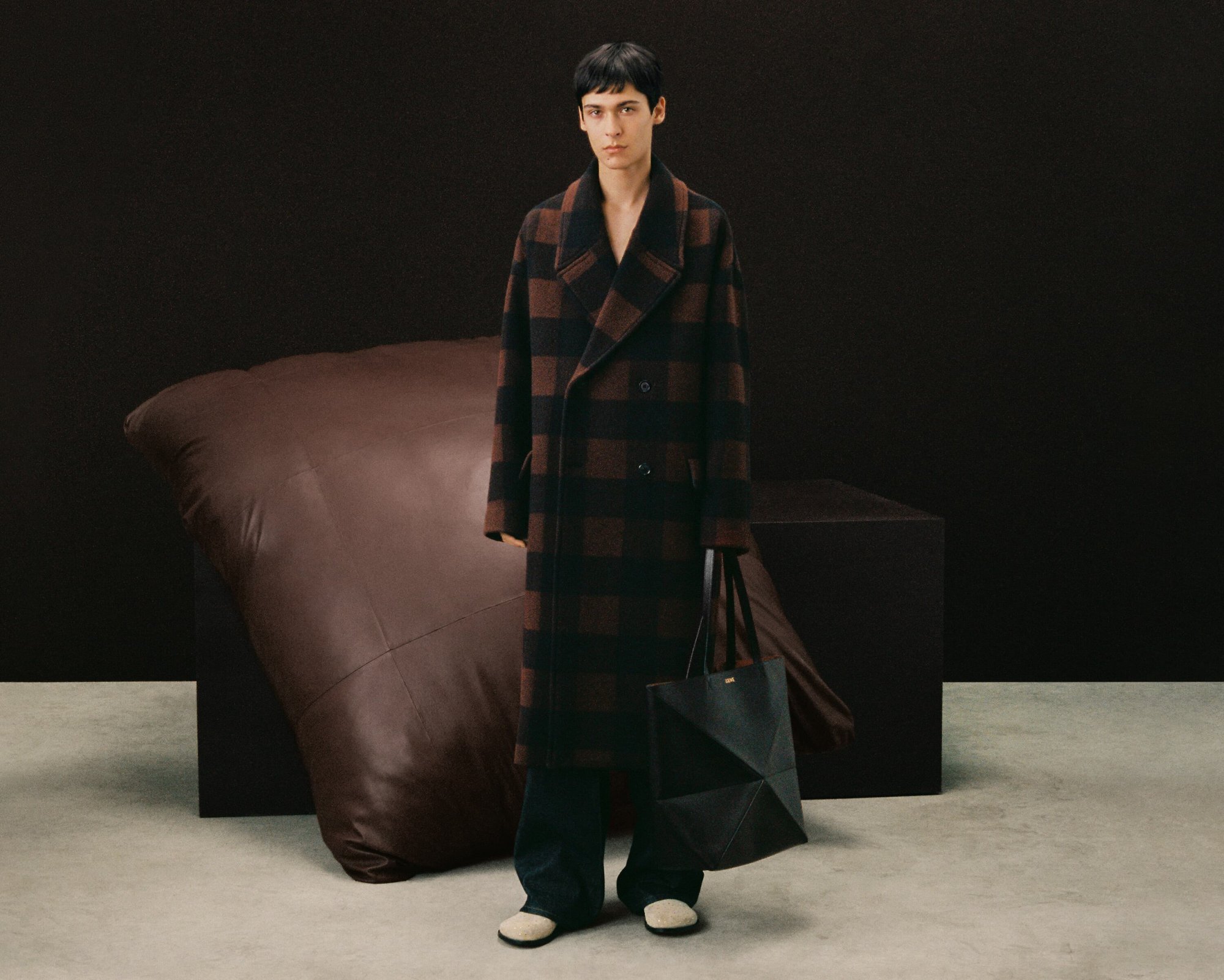 A young adult fashion model poses indoors in a brown and black checkerboard coat, dark trousers, and a geometric handbag.