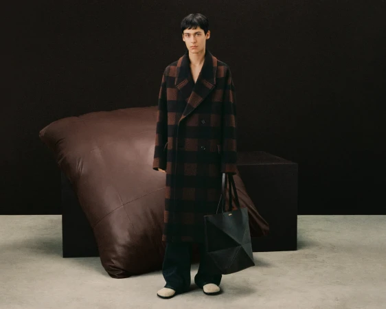 A young adult fashion model poses indoors in a brown and black checkerboard coat, dark trousers, and a geometric handbag.