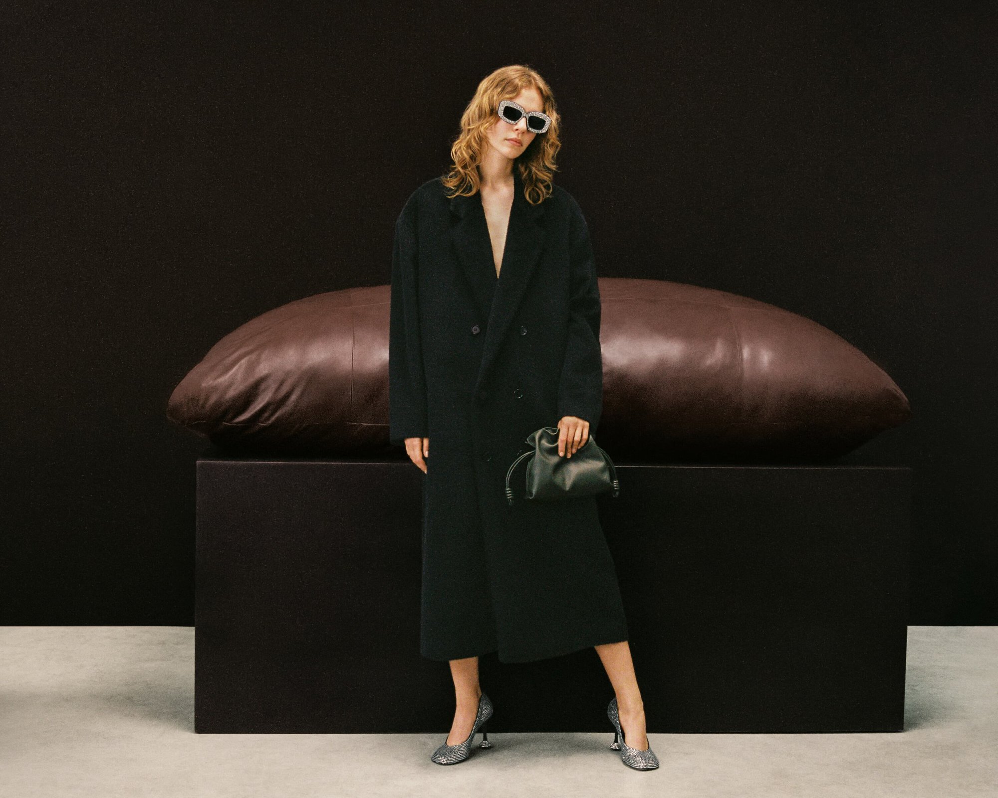 Model in dark coat and sunglasses standing before a large brown leather bolster on a dark rectangular plinth in a studio.