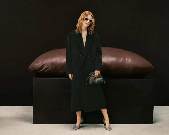 Model in dark coat and sunglasses standing before a large brown leather bolster on a dark rectangular plinth in a studio.
