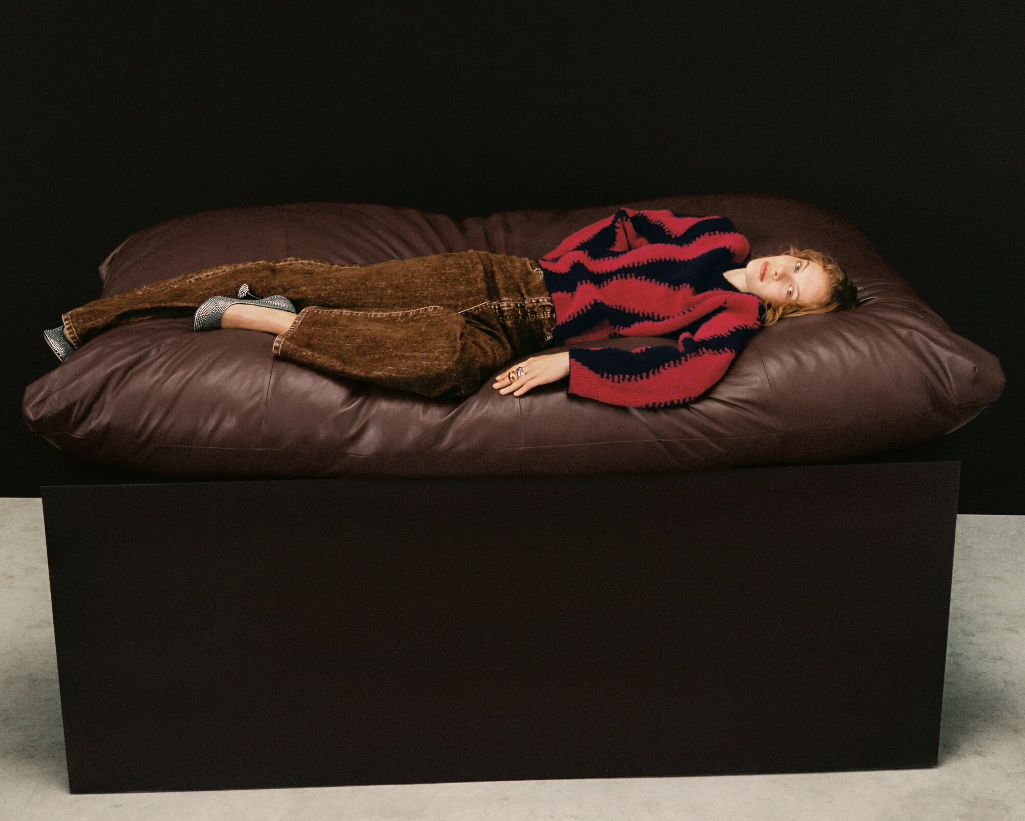 Woman lying on a brown leather couch in a photo studio. She is wearing brown pants and a red and black sweater.
