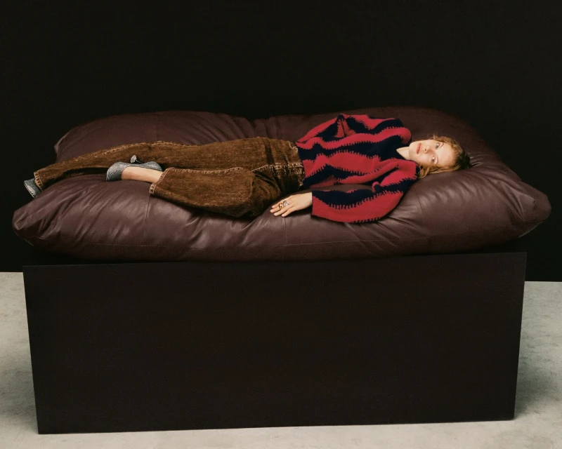Woman lying on a brown leather couch in a photo studio. She is wearing brown pants and a red and black sweater.