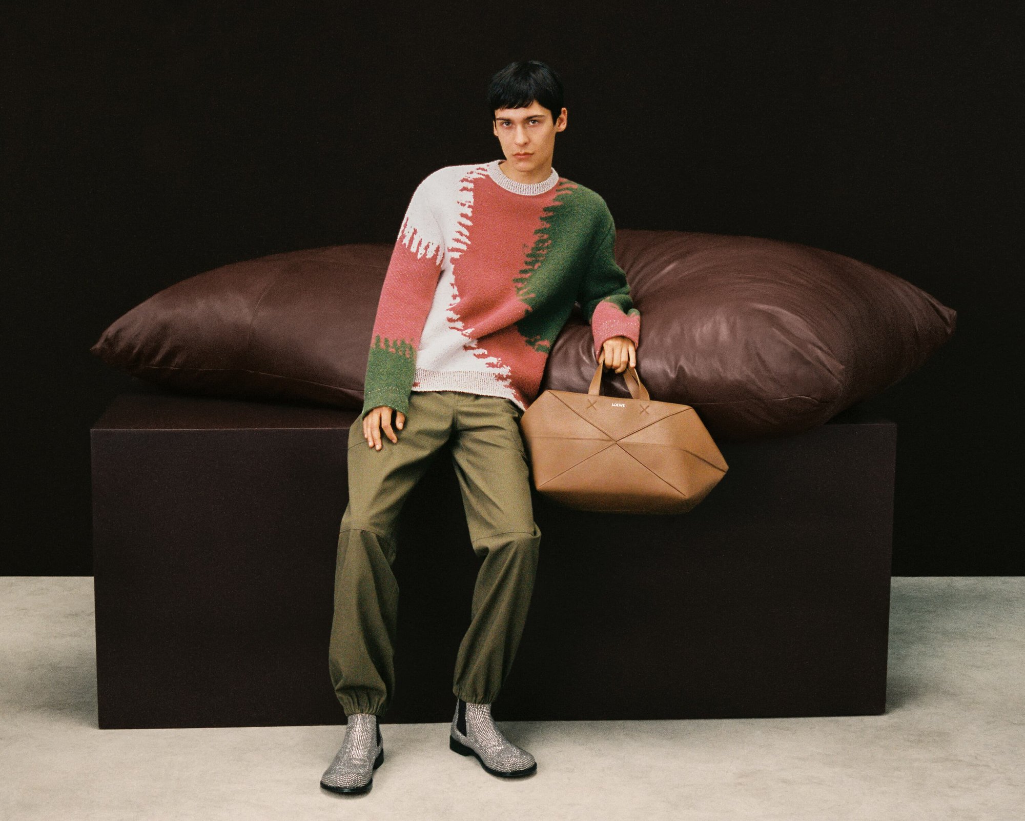 Fashion model poses in a colour-blocked sweater with a leather bag, green trousers, and metallic boots against a dark backdrop.