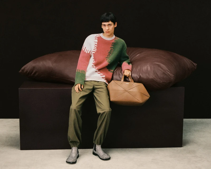 Fashion model poses in a colour-blocked sweater with a leather bag, green trousers, and metallic boots against a dark backdrop.