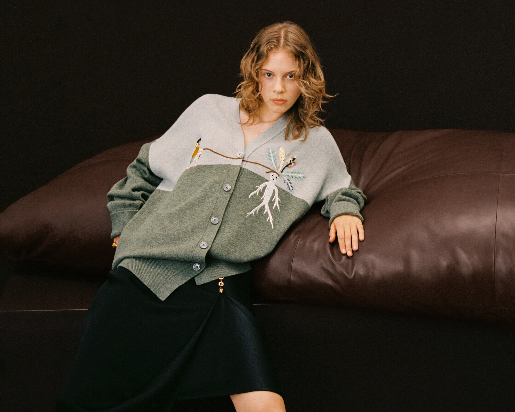 Adult woman poses in a two-tone cardigan with tree embroidery, against a dark backdrop with a brown leather couch.