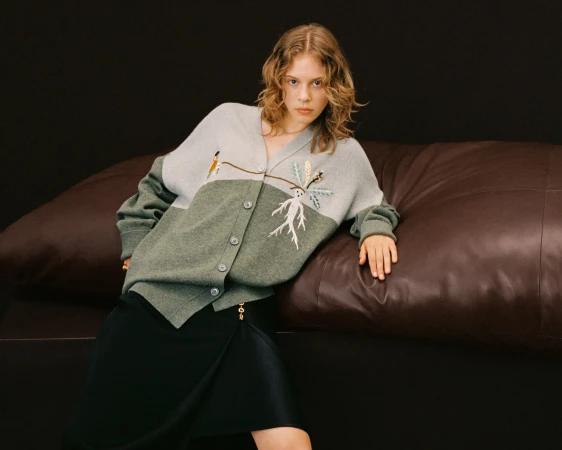 Adult woman poses in a two-tone cardigan with tree embroidery, against a dark backdrop with a brown leather couch.