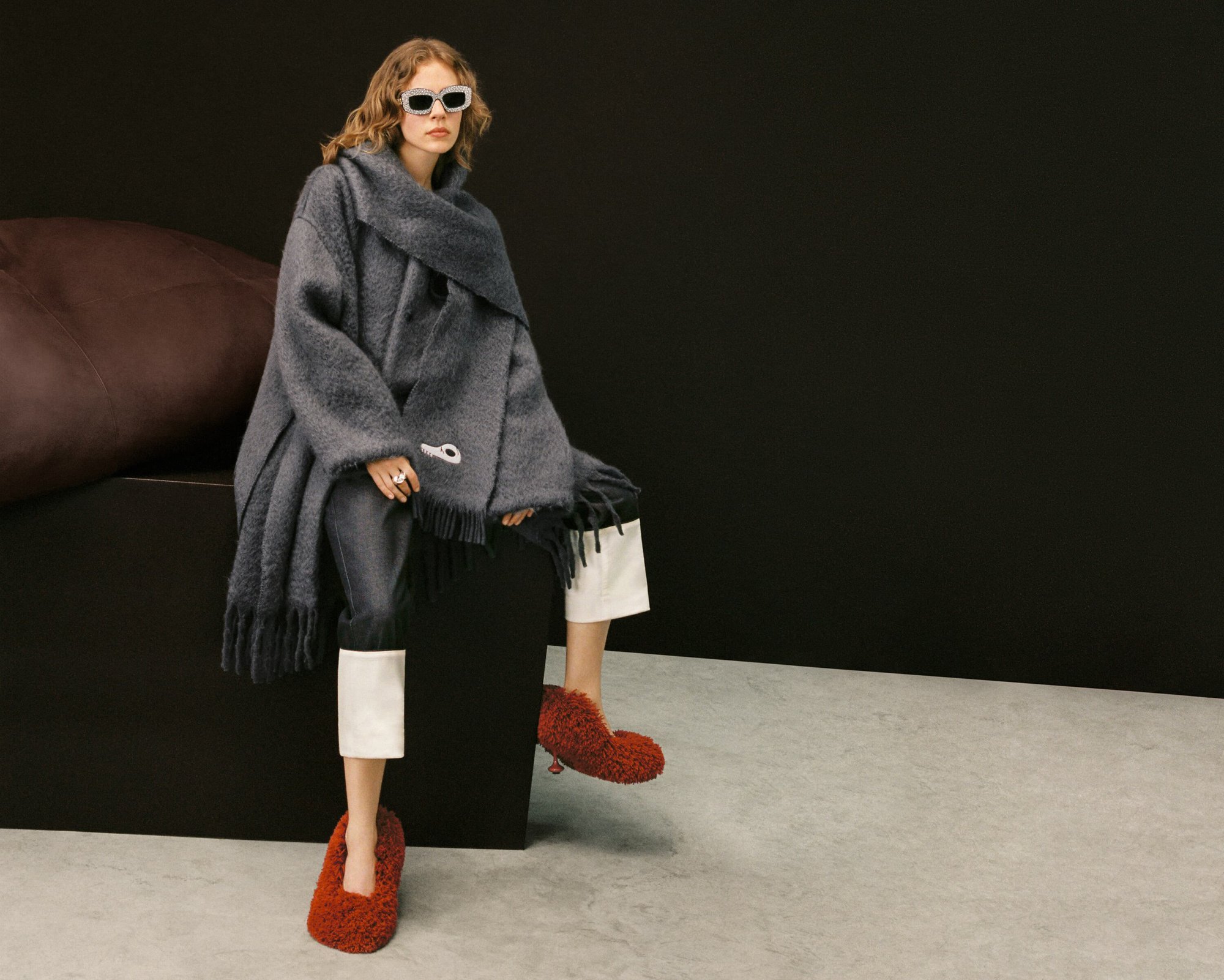 A woman sits on a black cube wearing sunglasses, a gray wool shawl, gray pants, white socks, and red fuzzy shoes in a studio.